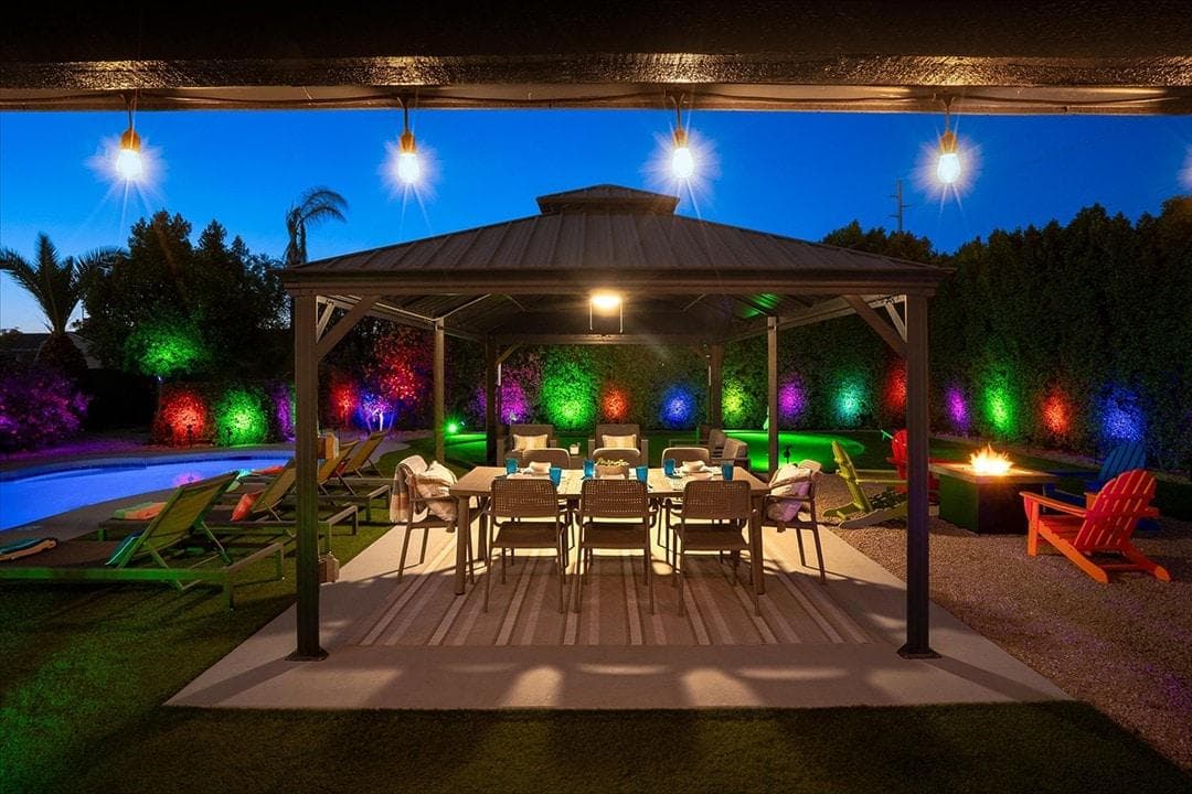 Gazebo, heated pool, lounge chairs, fire pit, dining table and chairs, and putting green at night lit up by string lights and colored LED solar lighting