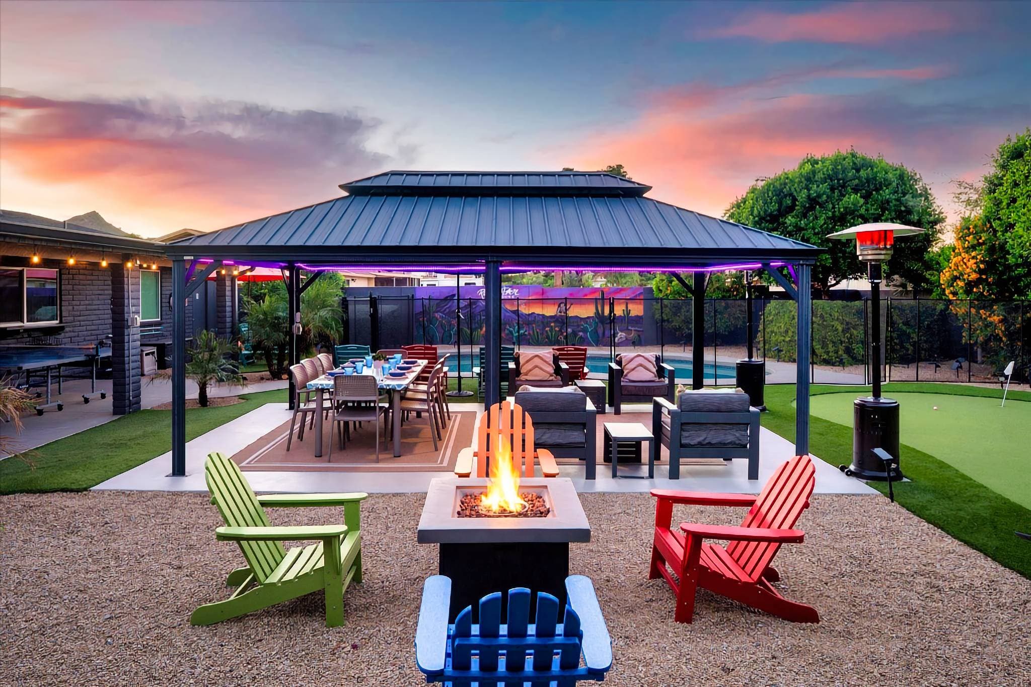 Evenings glow around the fire pit, where colorful Adirondack chairs, a covered dining space, and cozy lounge seating create the perfect hangout. With a putting green, heaters, and LED lighting, every night feels like a private resort experience.
